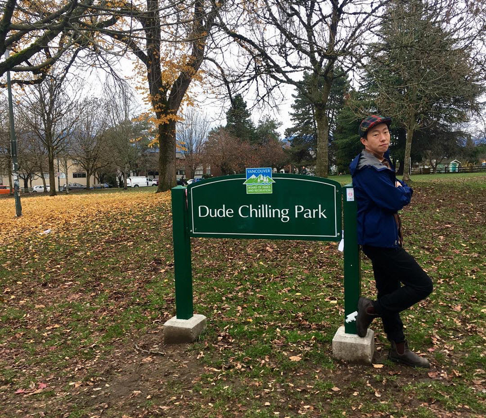 A photo of William Hsu. He's wearing dark clothes and a hat, leaning against a park sign that says "Dude Chilling Park." The backdrop shows some mostly bare trees with yellow leaves on the ground.