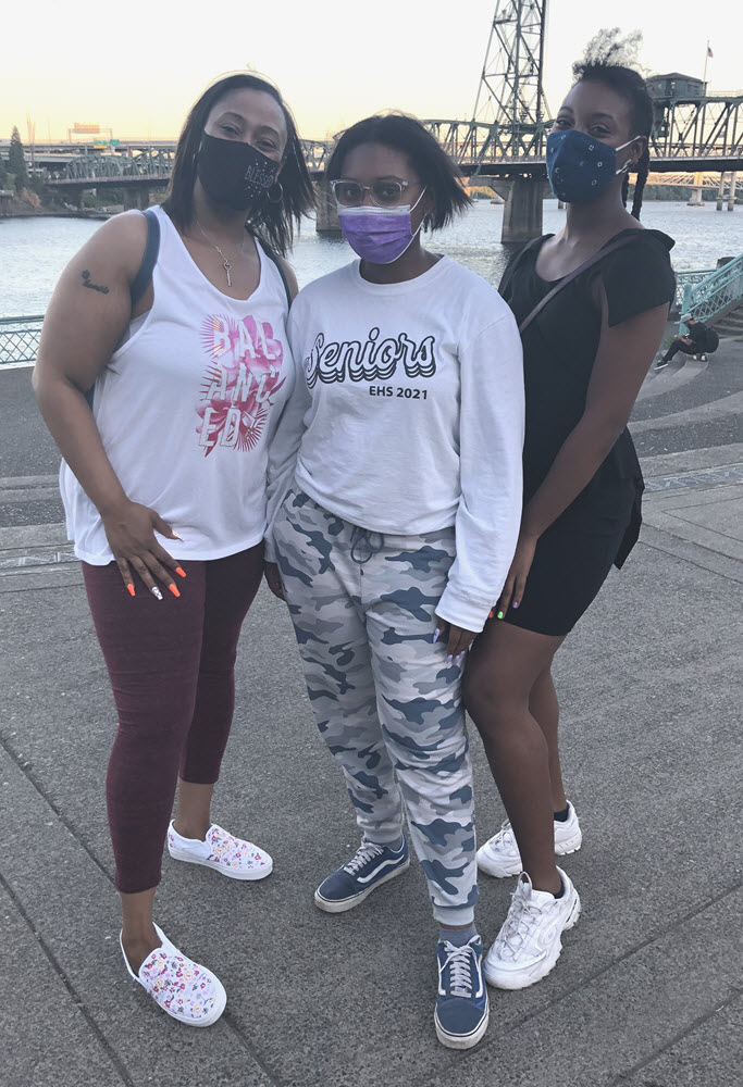 Three Black women in face masks pose on the Portland waterfront with a river and bridge in the background