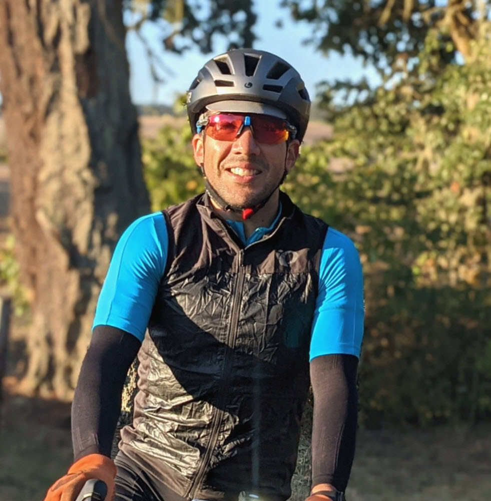 Ramon faces the camera, smiling. He's wearing a bike helmet, sunglasses, and a turquoise cycling jersey over black long sleeves, with a black vest, and orange gloves. His hand is visibly perched on top of bike handlebars.