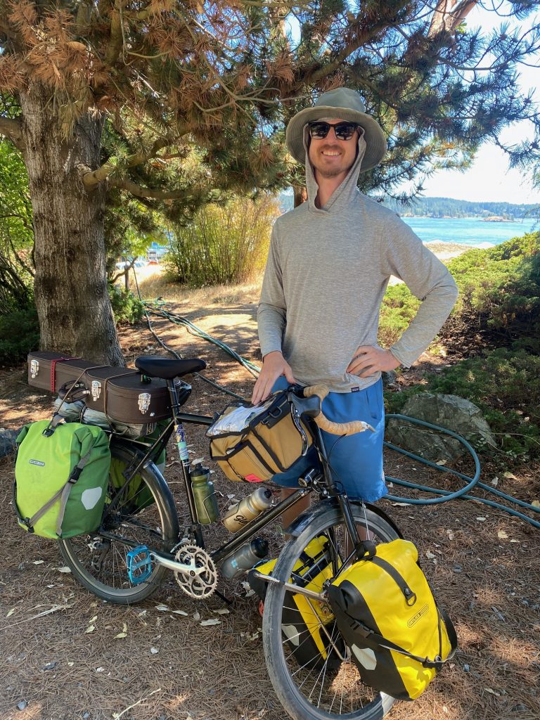 Guthrie stands in the shade with a distant view of water behind. He wears a hoodie, hat, and sunglasses, and stands next to his bike, loaded up with stuffed panniers.