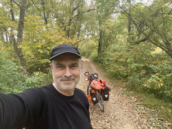 A man taking a selfie centered in the frame on a dirt path in green woods. He's wearing a black shirt and hat and smiling. Behind him, his bike is leaning and covered with panniers and other equipment.