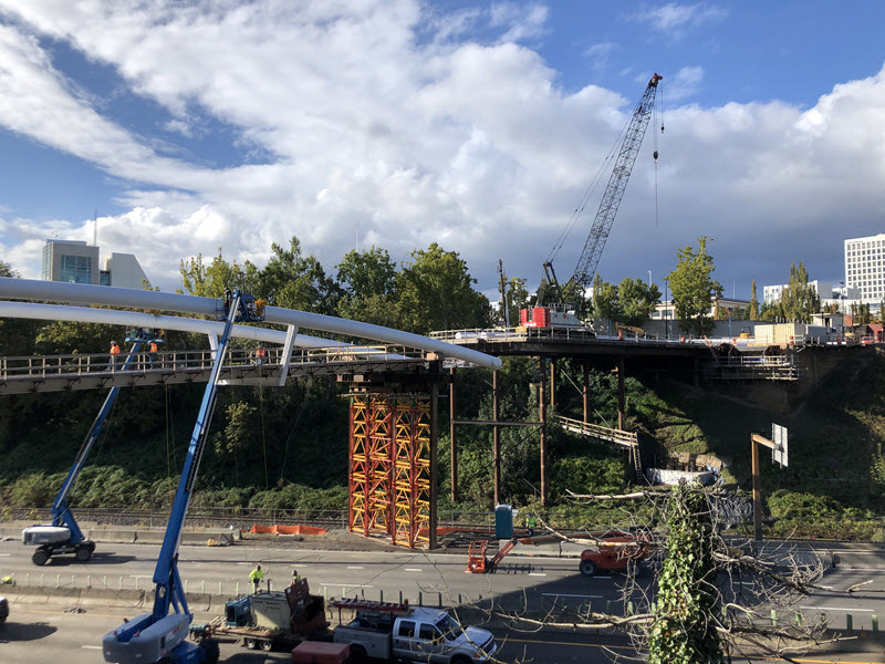 Construction site on highway, with cranes on the highway and platform above, showing part of new bridge as it's being installed. Backdrop shows blue sky with fluffy white clouds. 