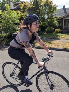 Sofie wears a gray shirt and black pants, sunglasses, and a black bike helmet. She rides her bike, smiling, down a neighborhood street.