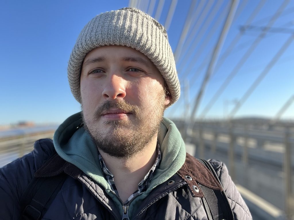 Close up selfie of Iain looking just off camera. He has a beanie and beard, light skin, and is wearing a sweatshirt and jacket. The blurred background shows bridge supports. 